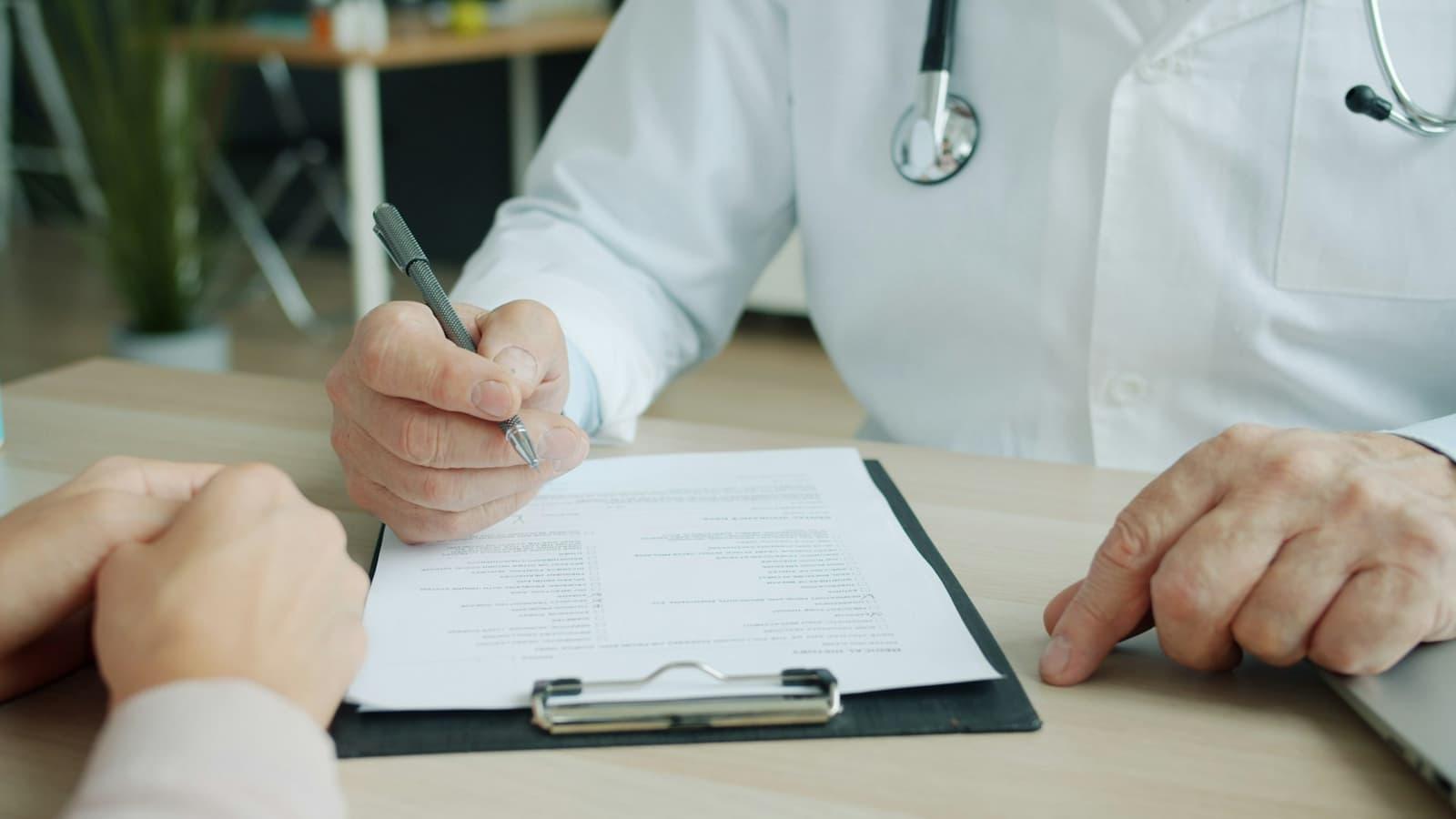 A clinician completing a form on a clipboard across a desk from a patient.