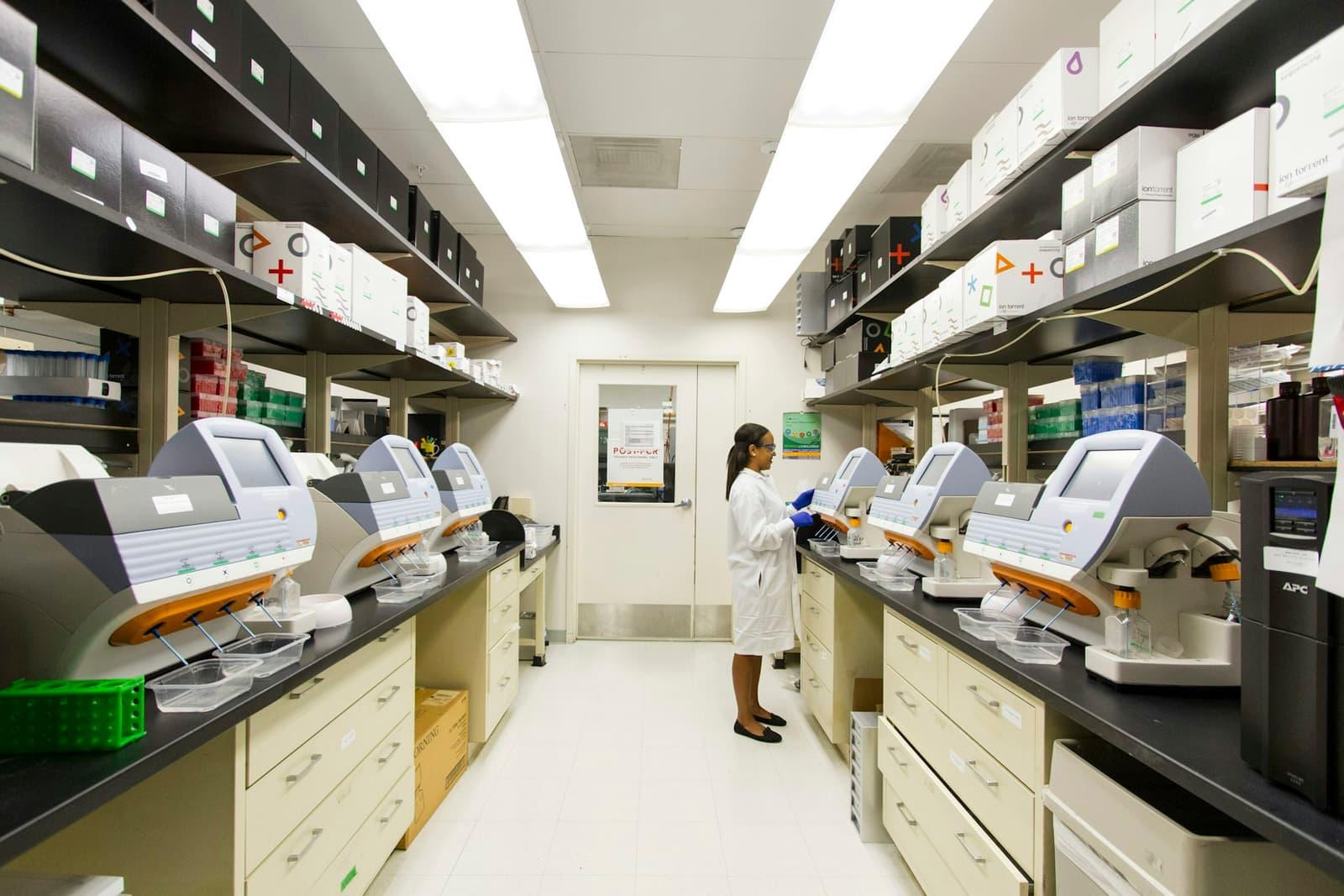 A hospital laboratory with rows of automated diagnostic analysers and a technician at work.