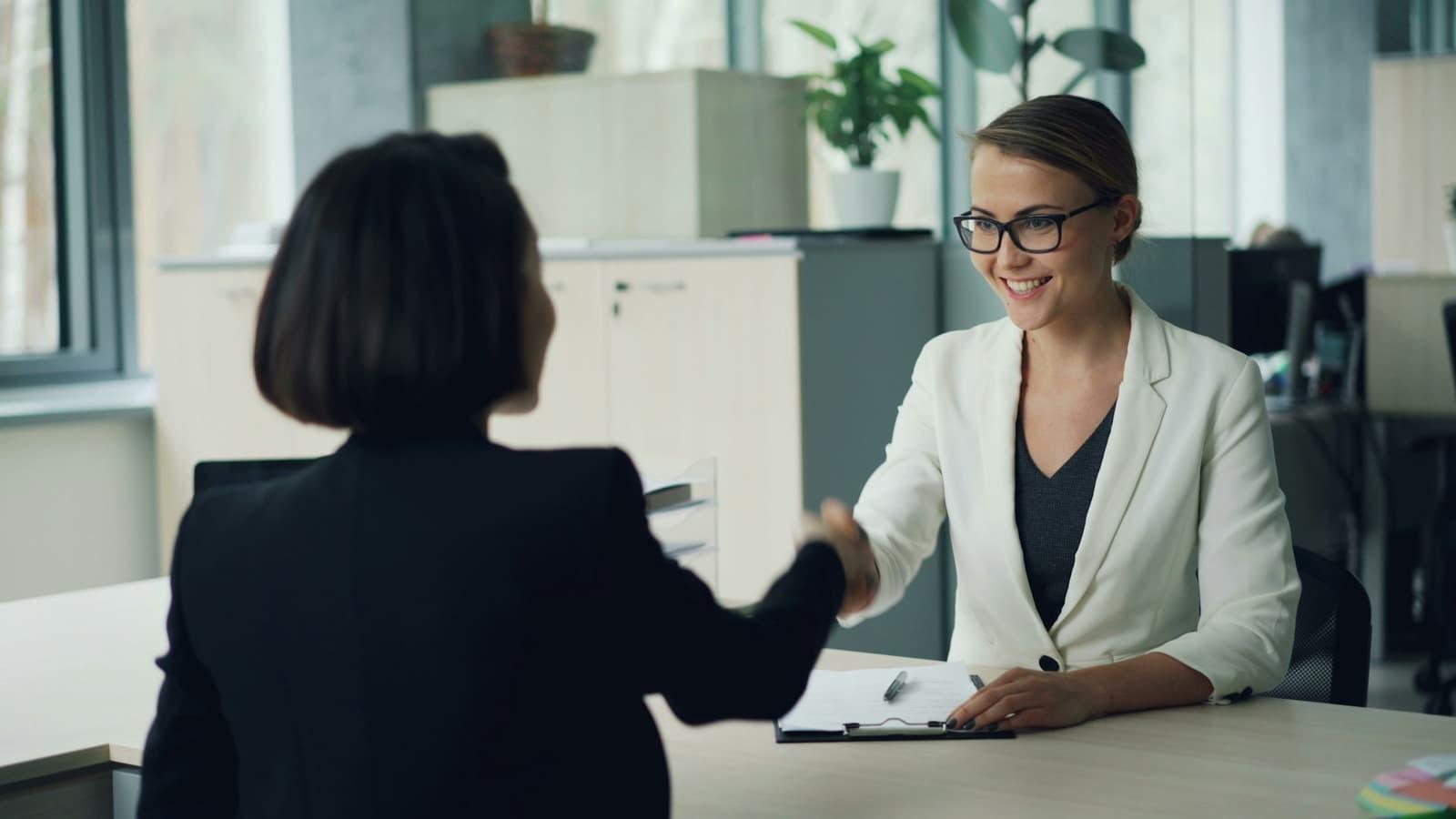 Two professionals shaking hands across a desk at the close of a meeting.